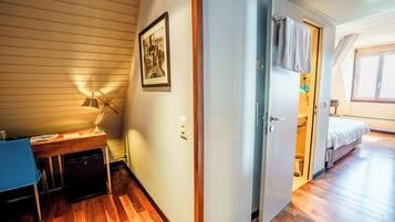 A room with a wooden desk, a lamp, a framed picture, and a bed in the adjacent room.