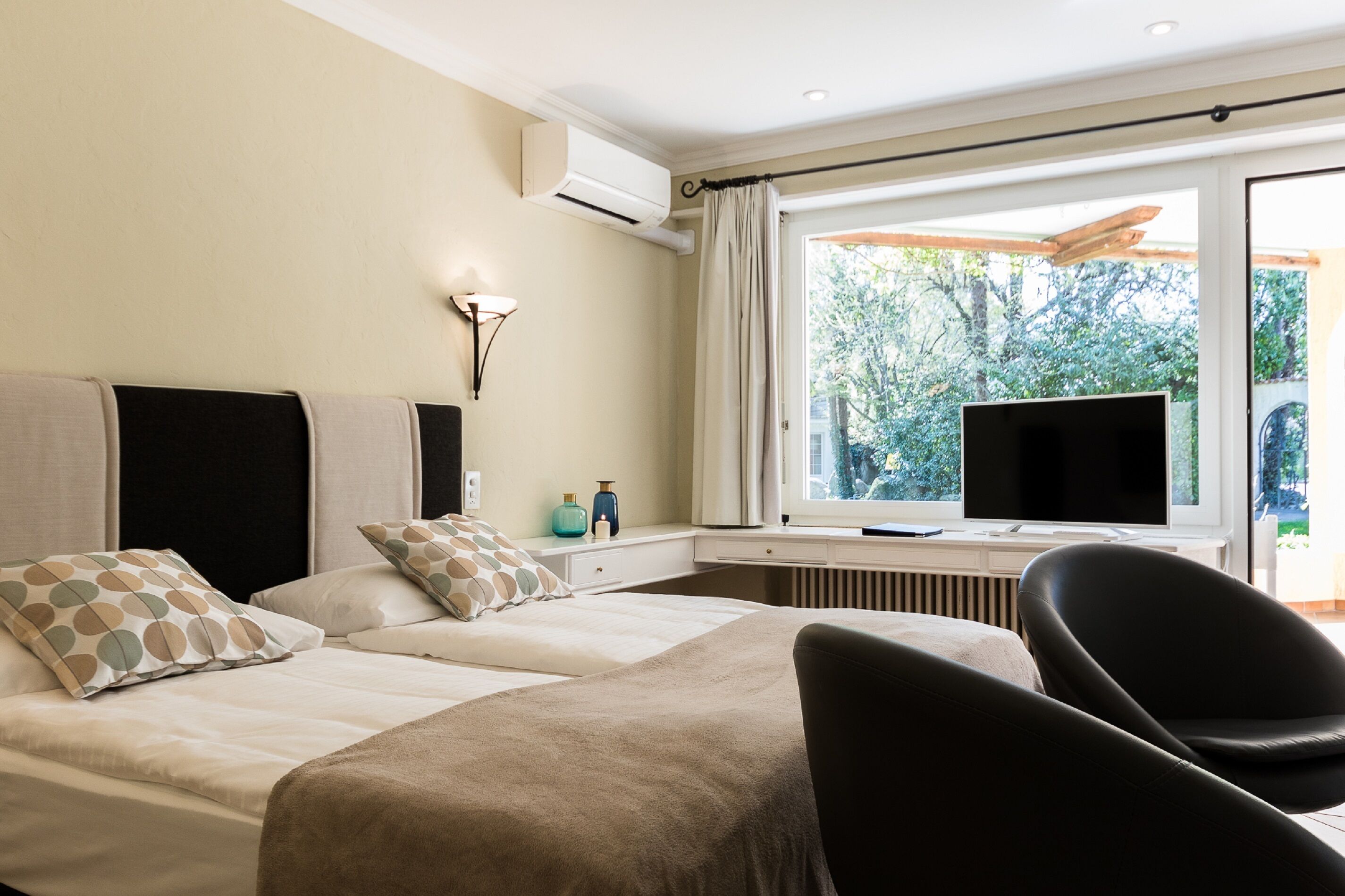 A hotel room with two beds, a TV, and a window with a view of greenery.