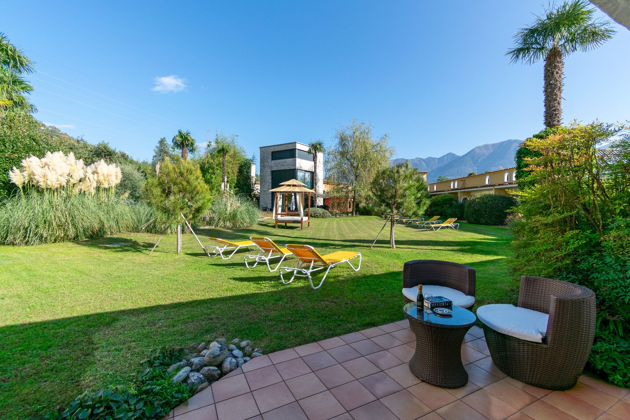 Eine Terrassenfläche mit Gartenmöbeln, einem kleinen Tisch und Blick auf ein modernes Gebäude sowie Berge im Hintergrund.