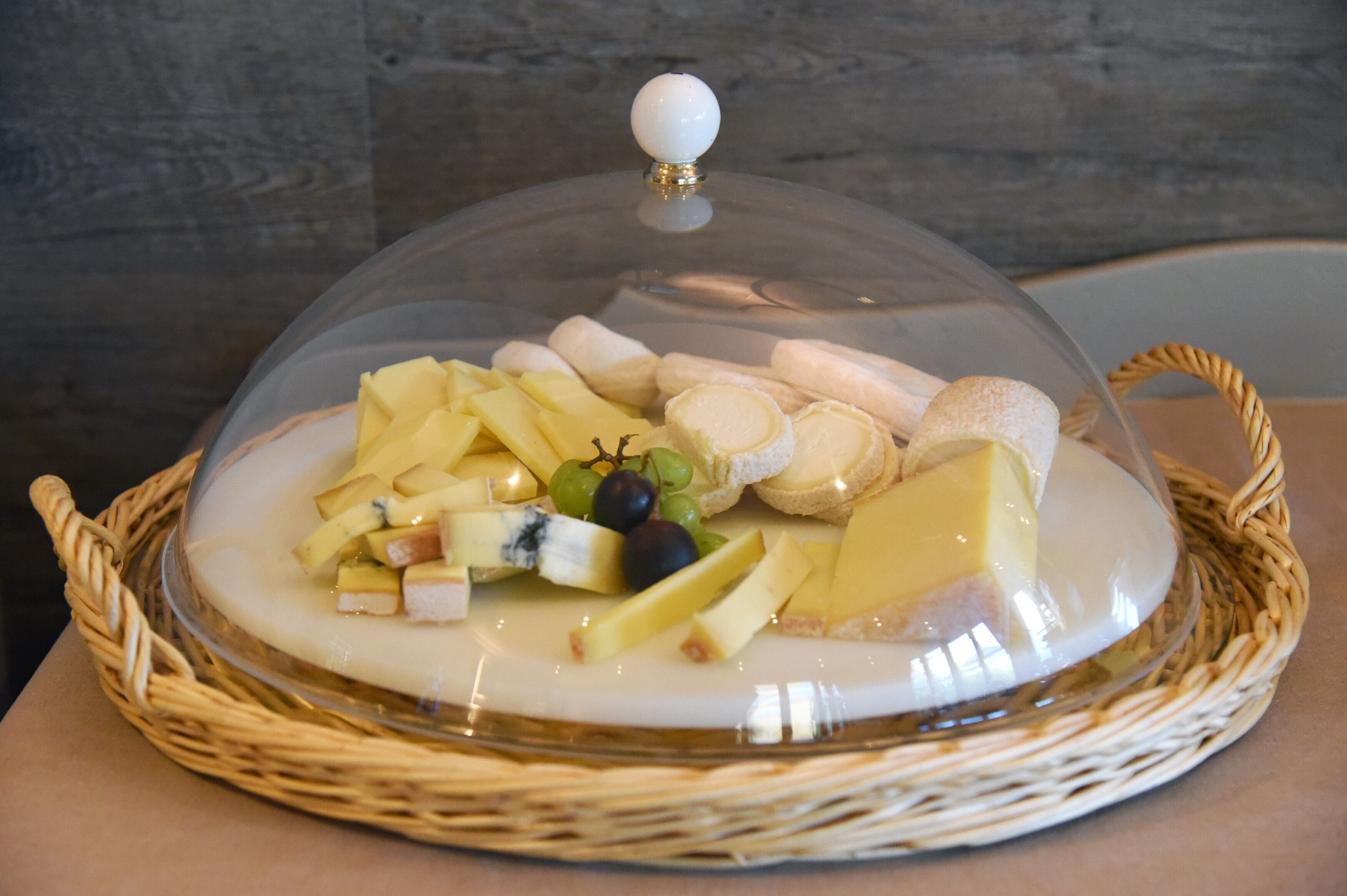 Een fruitschaal met kaas, afgedekt met glas, op een rieten dienblad.