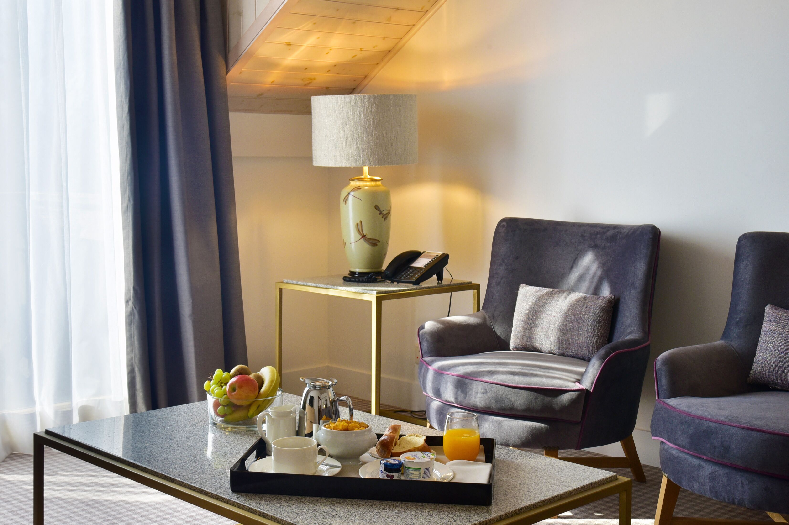 Ein Hotelzimmer mit Esstisch, gedecktem Frühstücksservice, zwei Sesseln, einer Lampe und einem Telefon auf einem Beistelltisch.