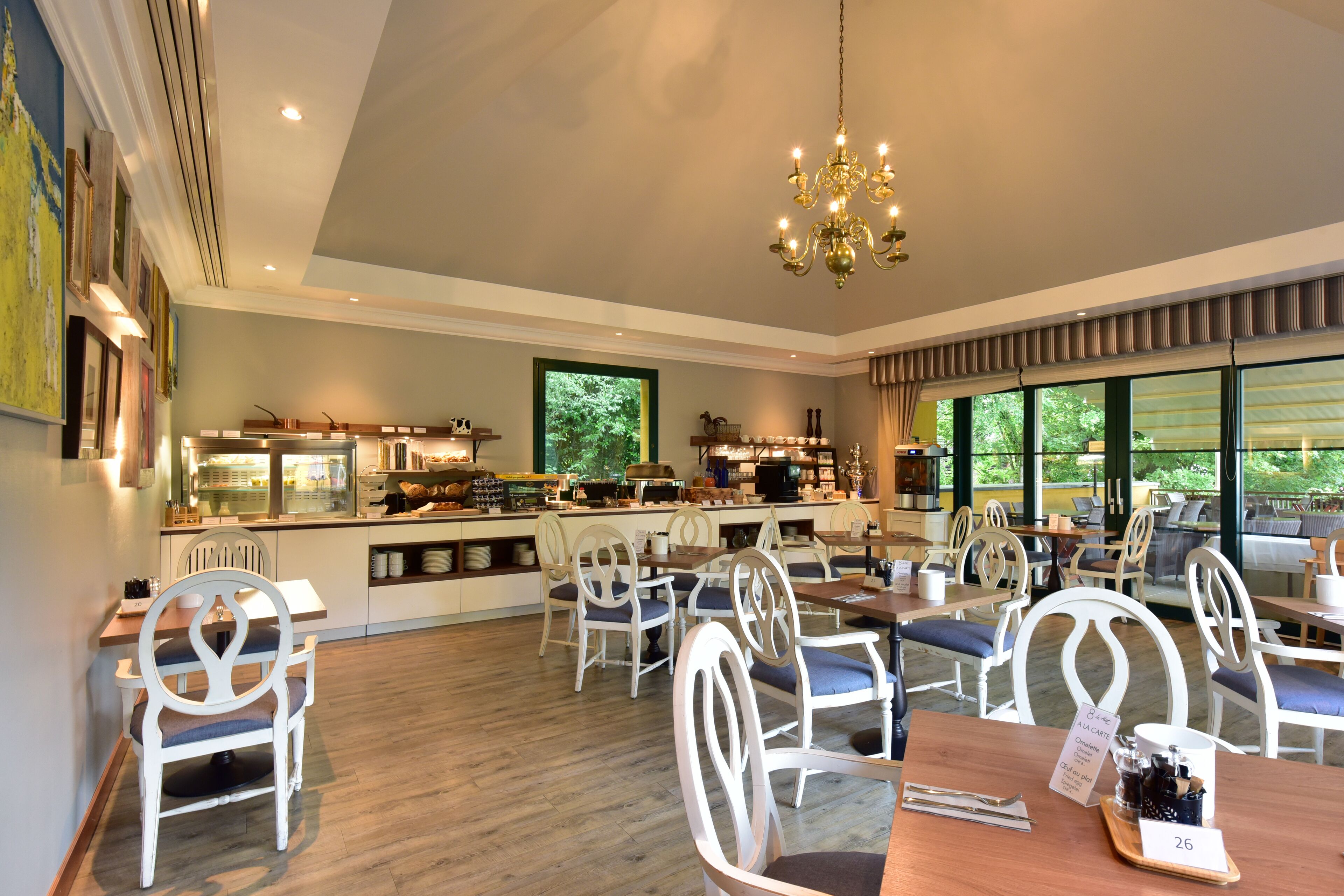 A spacious dining area with white chairs and tables, a chandelier, and a kitchen in the background.