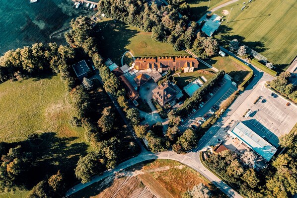 An aerial view of a property with a central building surrounded by trees, parking areas, and a small body of water. - Hotel La Barcarolle (Prangins)