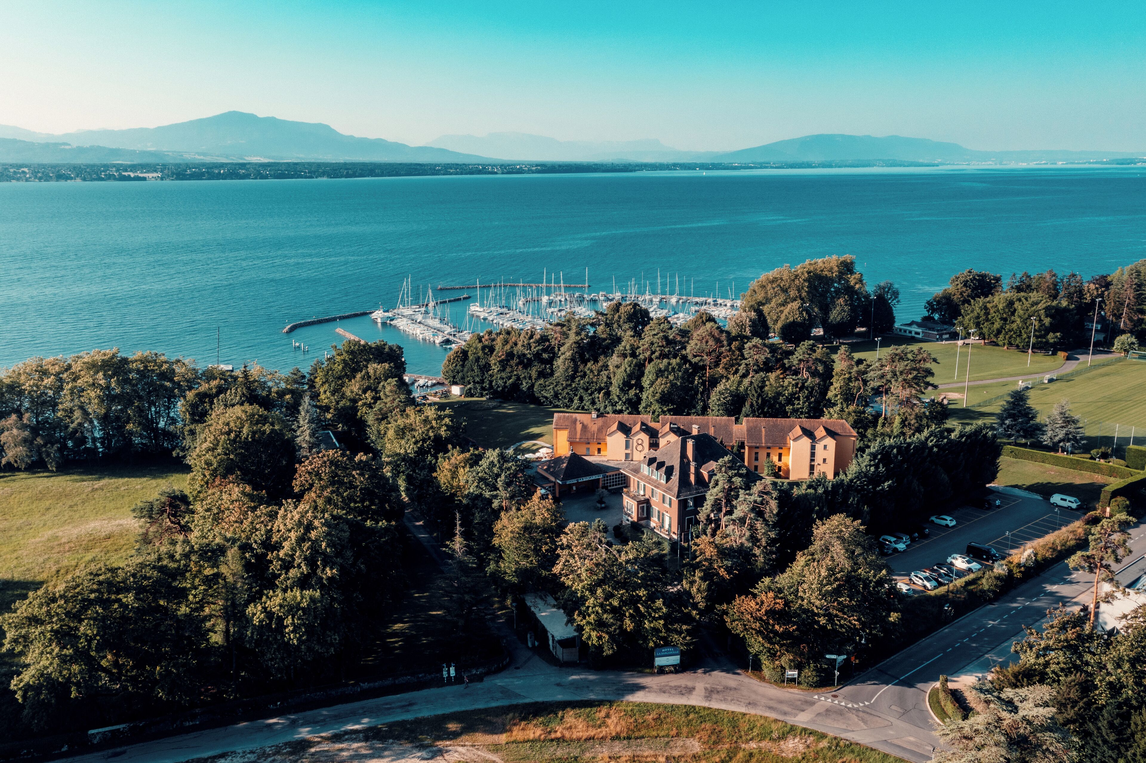 A large building surrounded by trees, a marina with boats, and a body of water with mountains in the background.