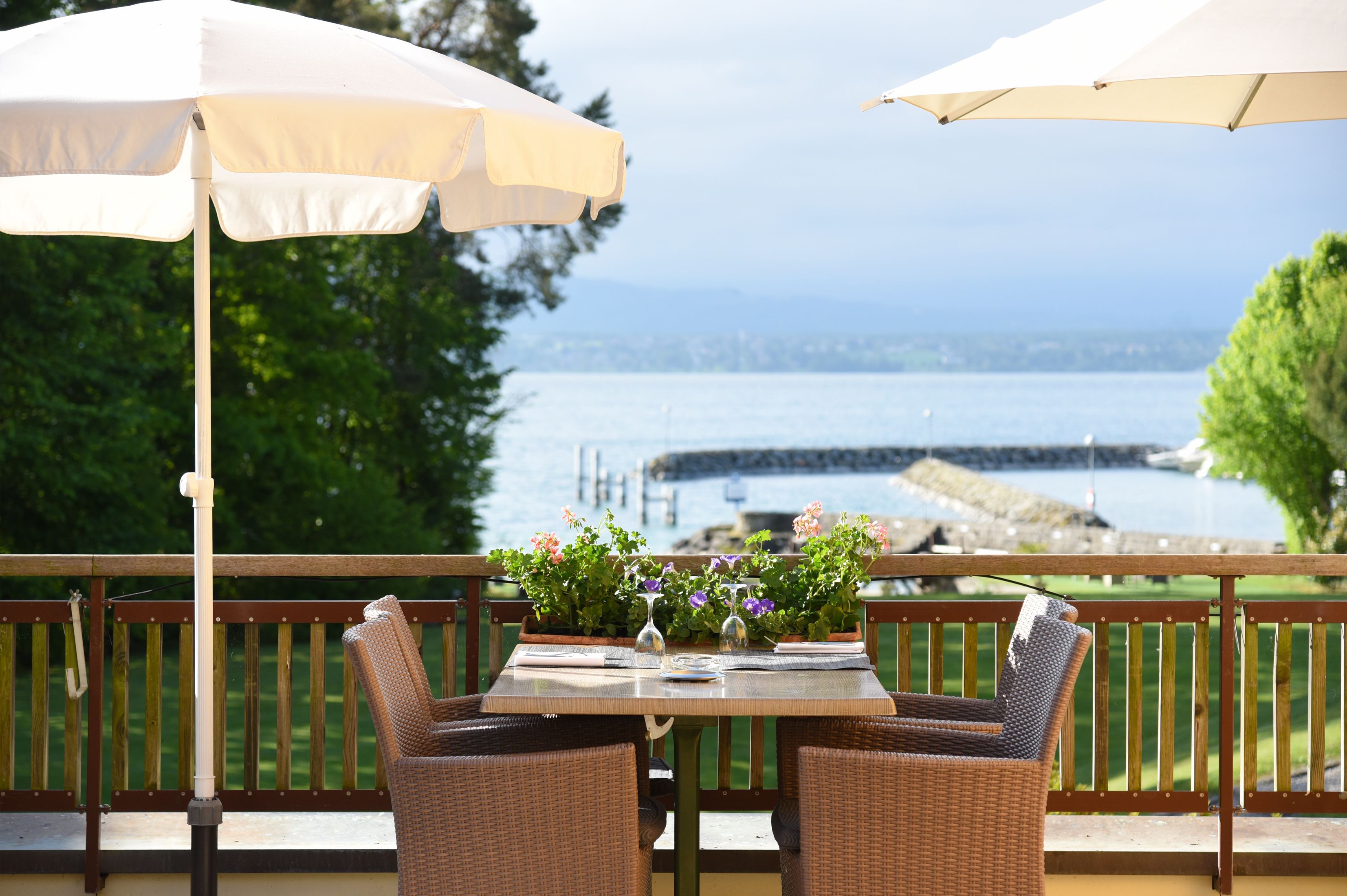 A table with two wicker chairs and a table setting, including a vase of flowers and a wine glass, on a balcony with a view of a body of water and a dock.