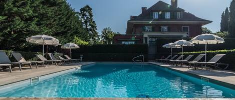 A swimming pool with lounge chairs and umbrellas, surrounded by a stone deck and a large house in the background.