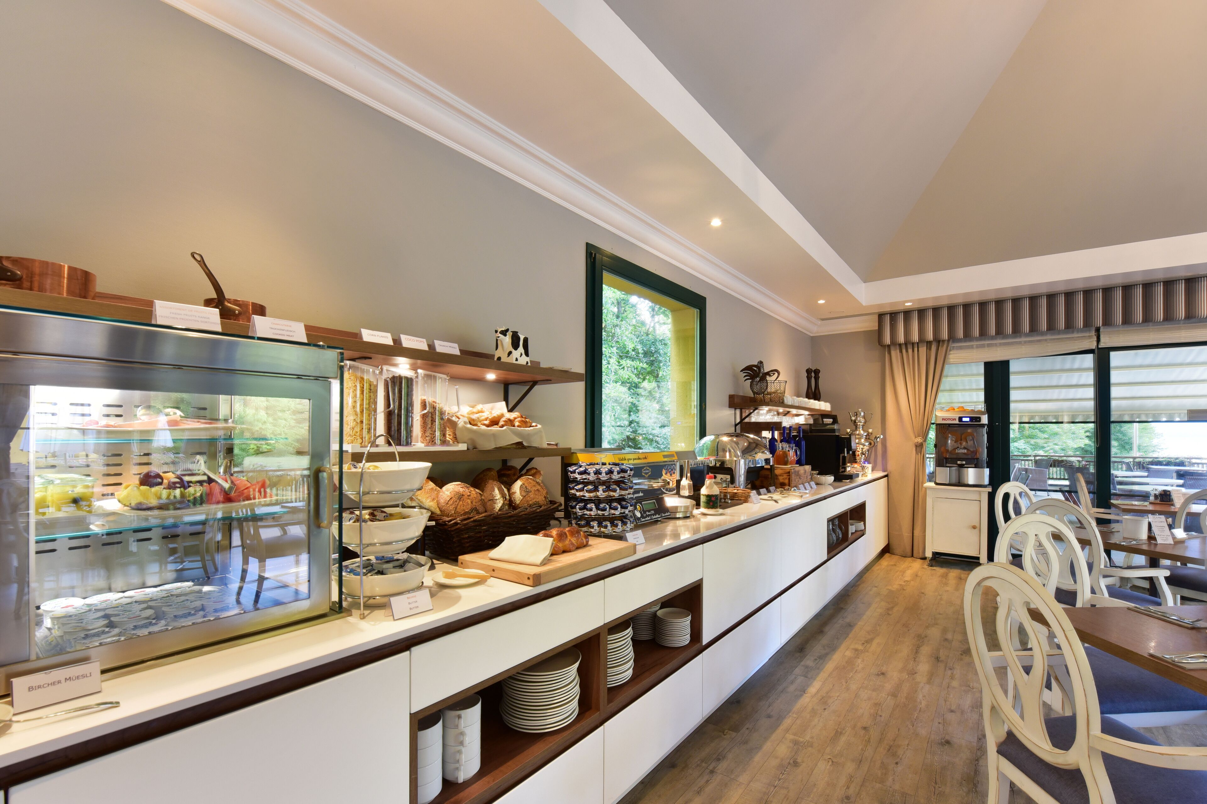 A breakfast buffet with various food items, a coffee station, and seating area with white chairs.