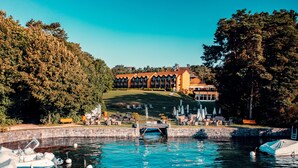 A lakeside hotel with a large building, a dock with boats, and a grassy area with outdoor seating. - Hotel La Barcarolle (Prangins)