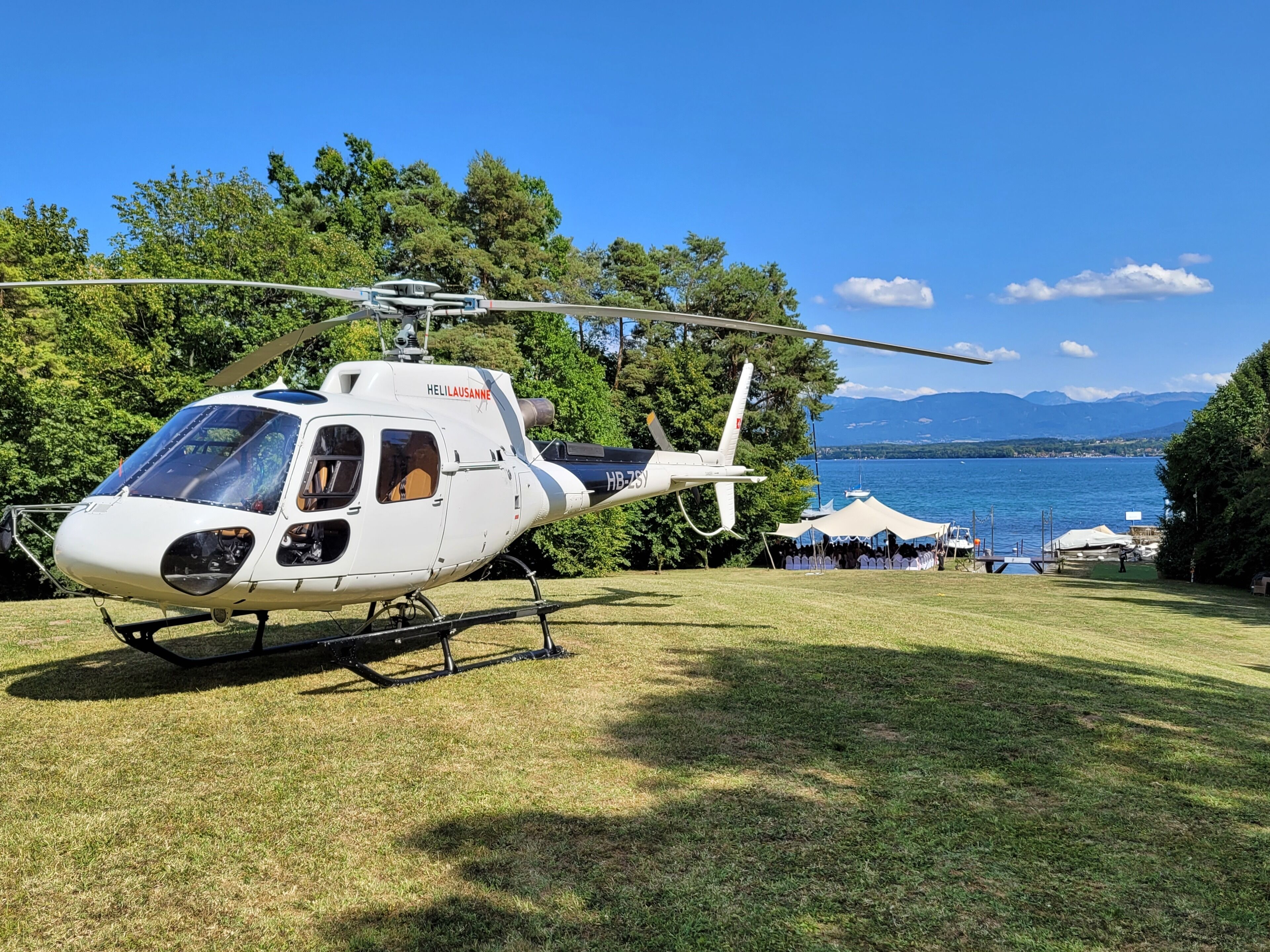 Um helicóptero branco com as marcações "Heli Lausanne" e "HB-ZYS" estacionado na grama perto de um lago, com tendas e pessoas ao fundo.