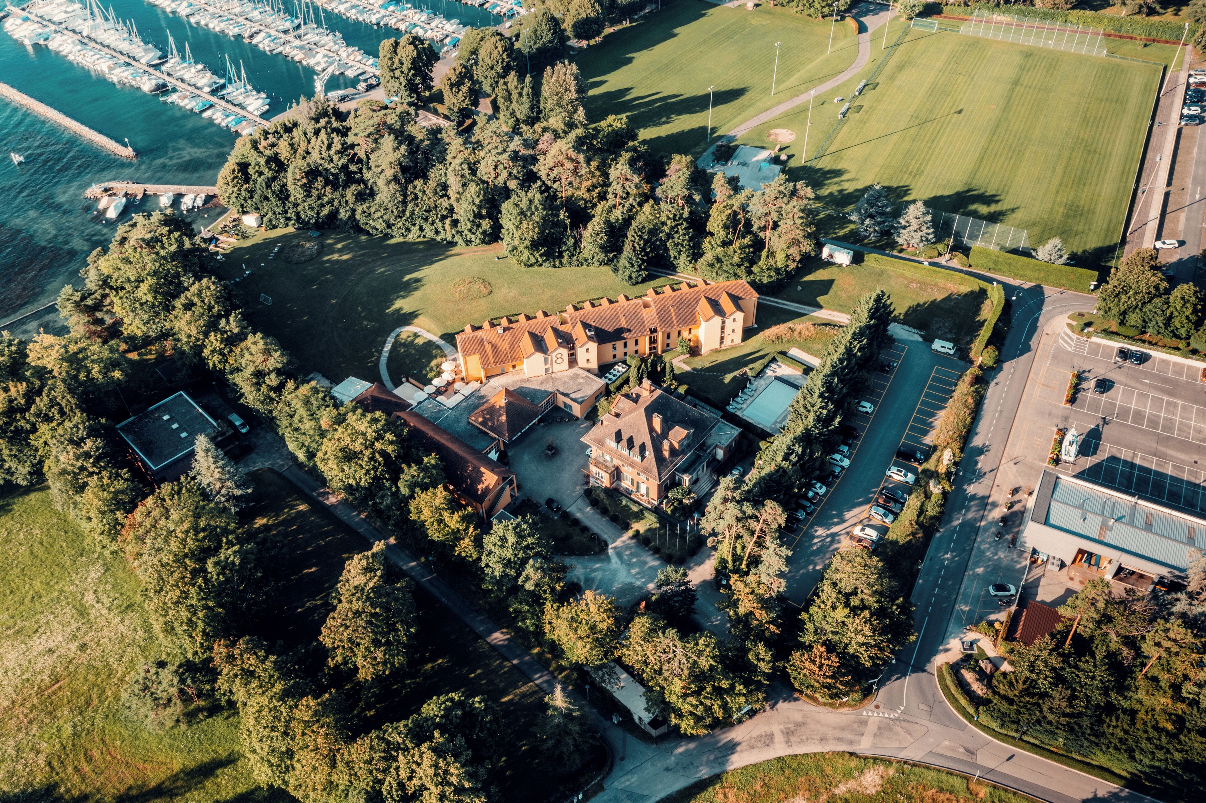 An aerial view of a large, historic building surrounded by dense trees, with a marina and a parking lot in the foreground.