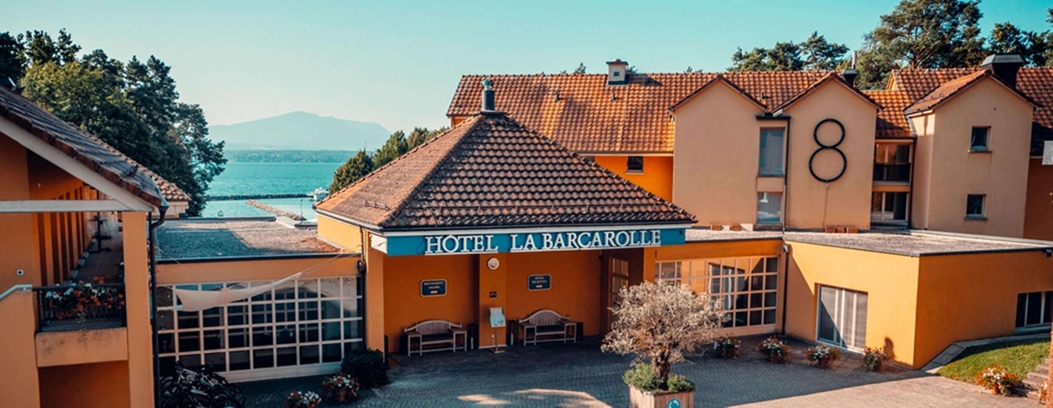 Un hotel con tejado de tejas, un cartel que dice "HOTEL LA BARCAROLLE" y vista al mar al fondo.