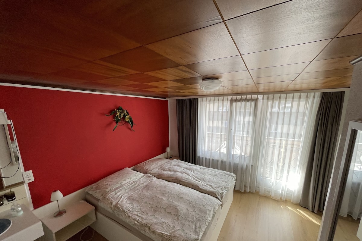 A bedroom with a wooden ceiling, a red wall, a bed with white bedding, a bedside table with a lamp, and a window with sheer curtains.