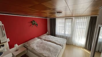 Un dormitori amb sostre de fusta, una paret vermella, un llit amb roba de llit blanca, una tauleta de nit amb una làmpada i una finestra amb cortines lleugeres.