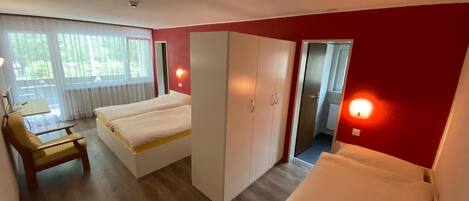 Quarto de hotel com duas camas, uma escrivaninha e uma cadeira. Há armários e uma porta que dá para outro cômodo.
