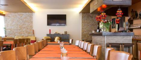 Una larga mesa de comedor con manteles anaranjados y sillas de madera, una pared de piedra y un televisor de pantalla plana montado en la pared.
