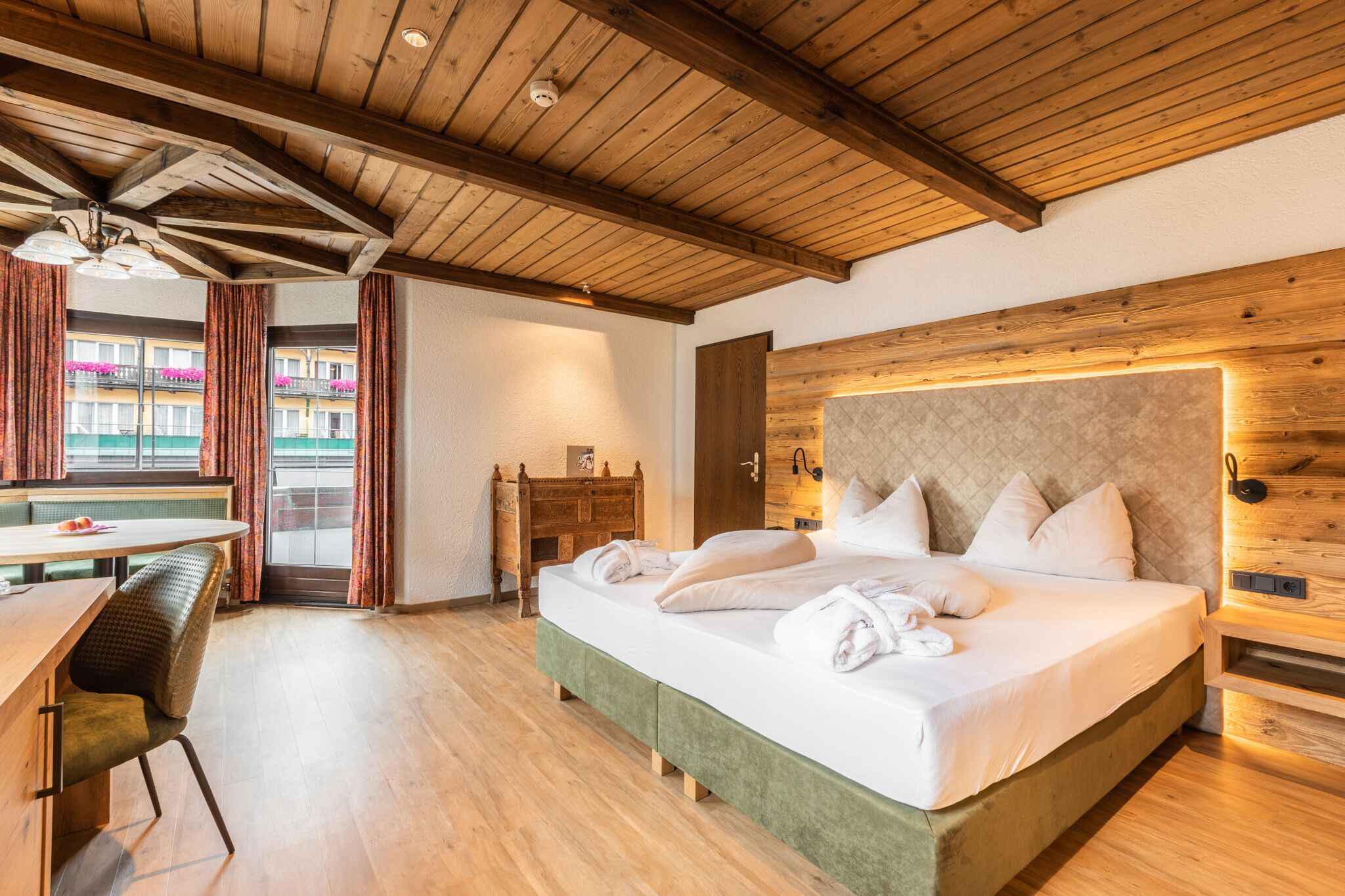 Camera d'albergo con soffitto in legno, un letto con lenzuola bianche, una scrivania in legno e vista su balconi fioriti.