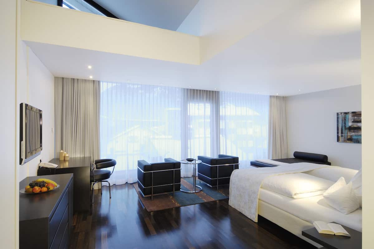 A modern hotel room with a bed, a desk with a telephone, a chair, a small table, and a sofa. There is a large window with curtains, and a painting on the wall.