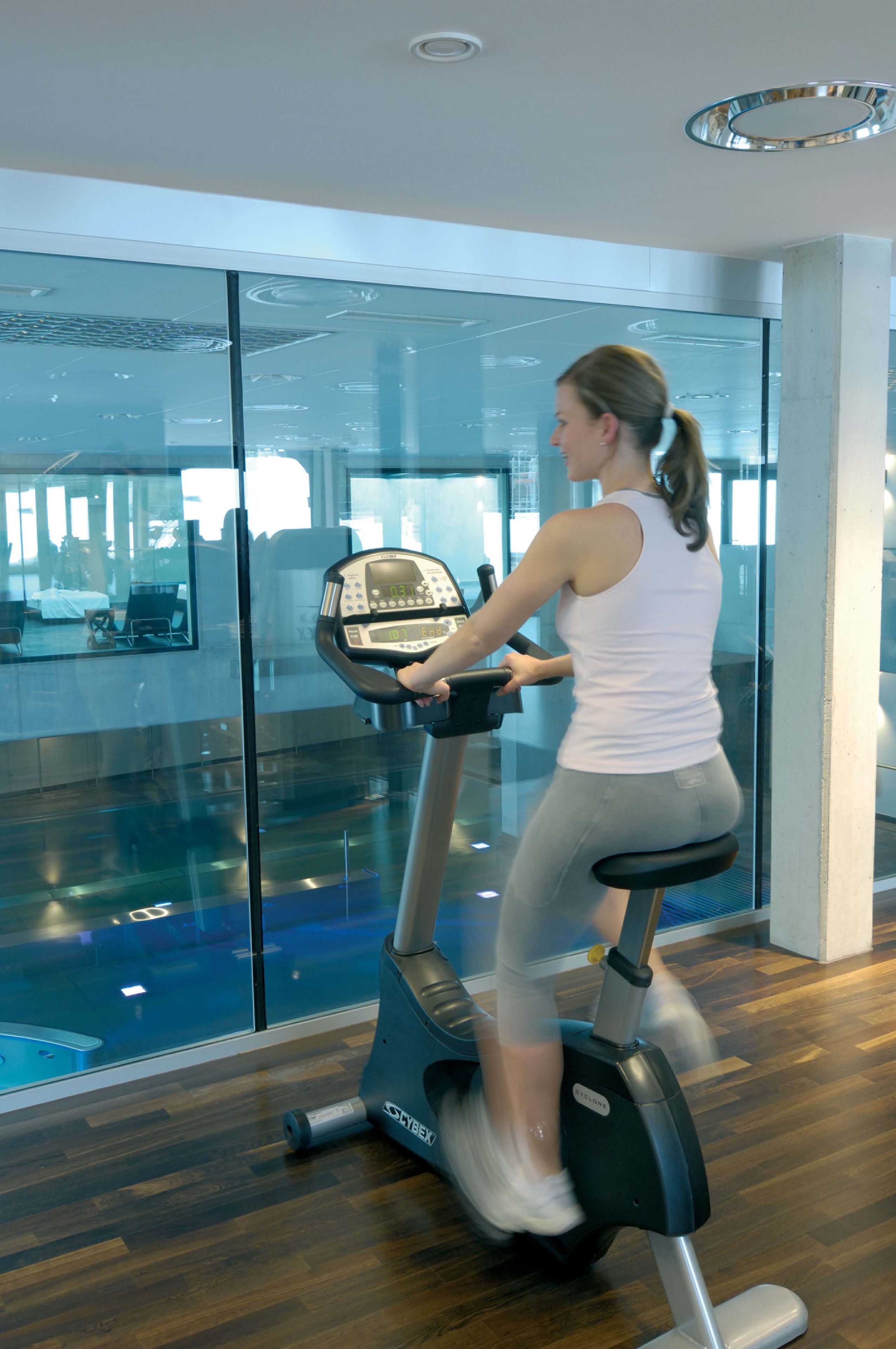 Une femme fait de l’exercice sur un vélo stationnaire dans une salle de sport moderne aux murs de verre et au plancher en bois.