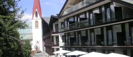 A modern hotel with a large glass facade, a red spire, and a rooftop lounge area with sun loungers and umbrellas.