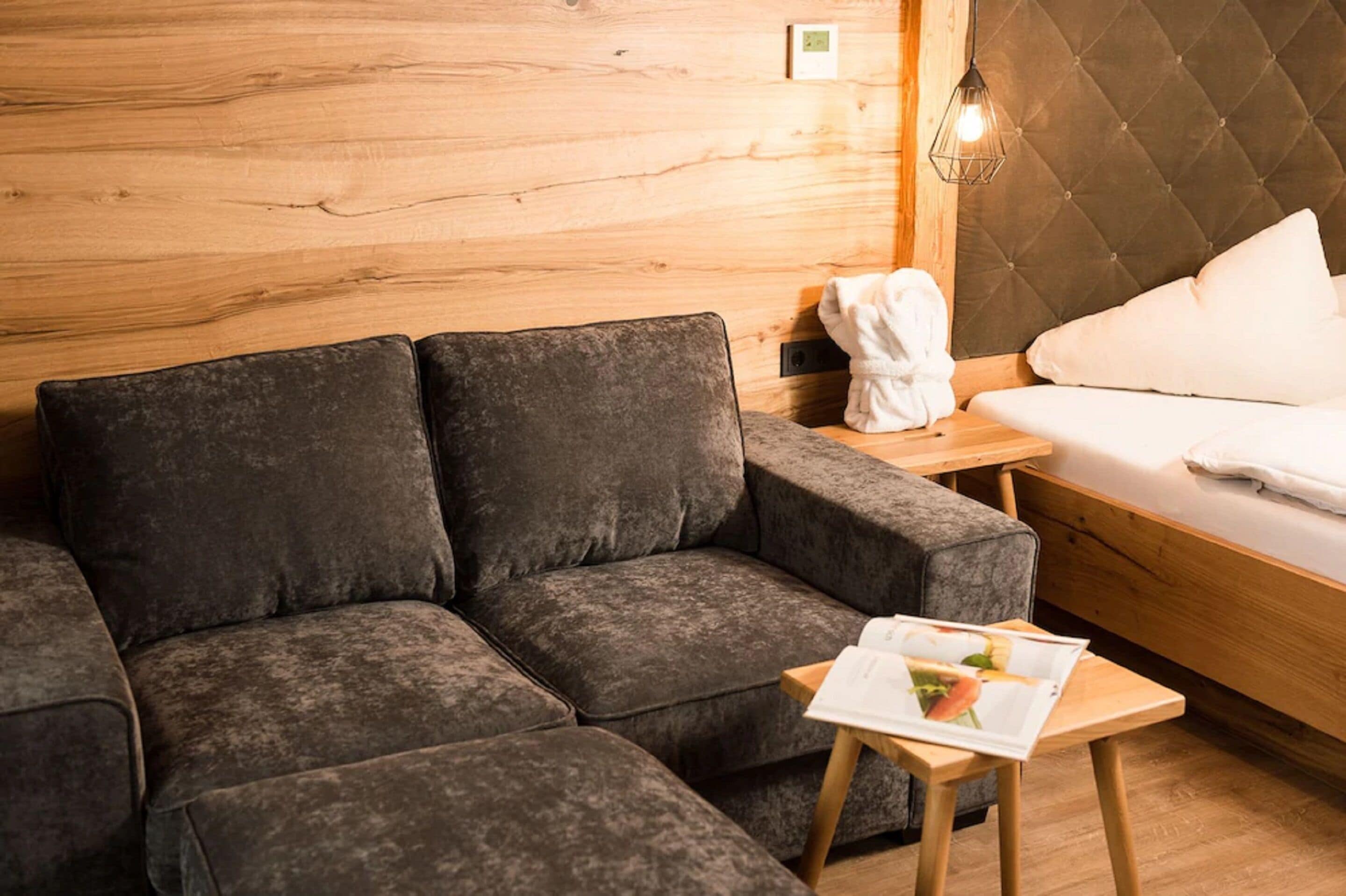 A modern room with a dark grey sofa, a wooden wall, a bed with white bedding, and a small table with a magazine.