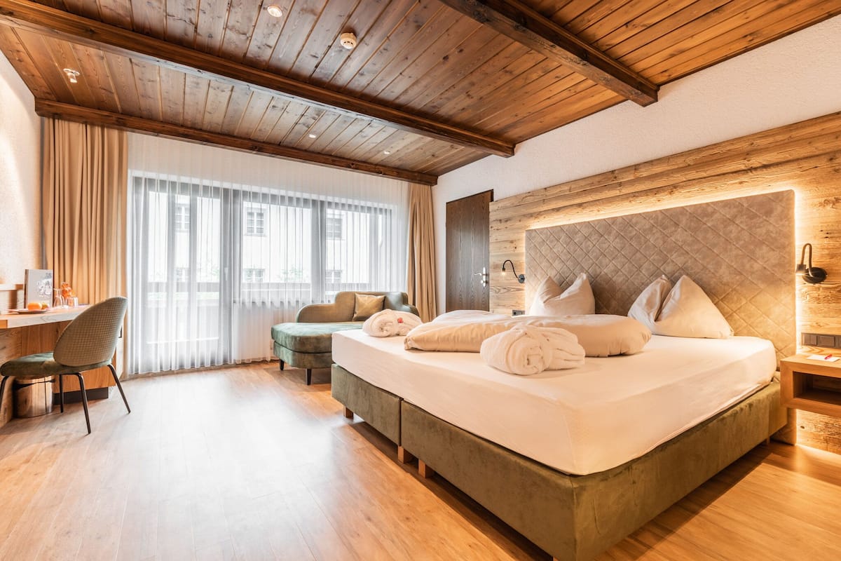 A hotel room with a wooden ceiling, a large bed with pillows and a blanket, a desk with a chair, and a window with curtains.