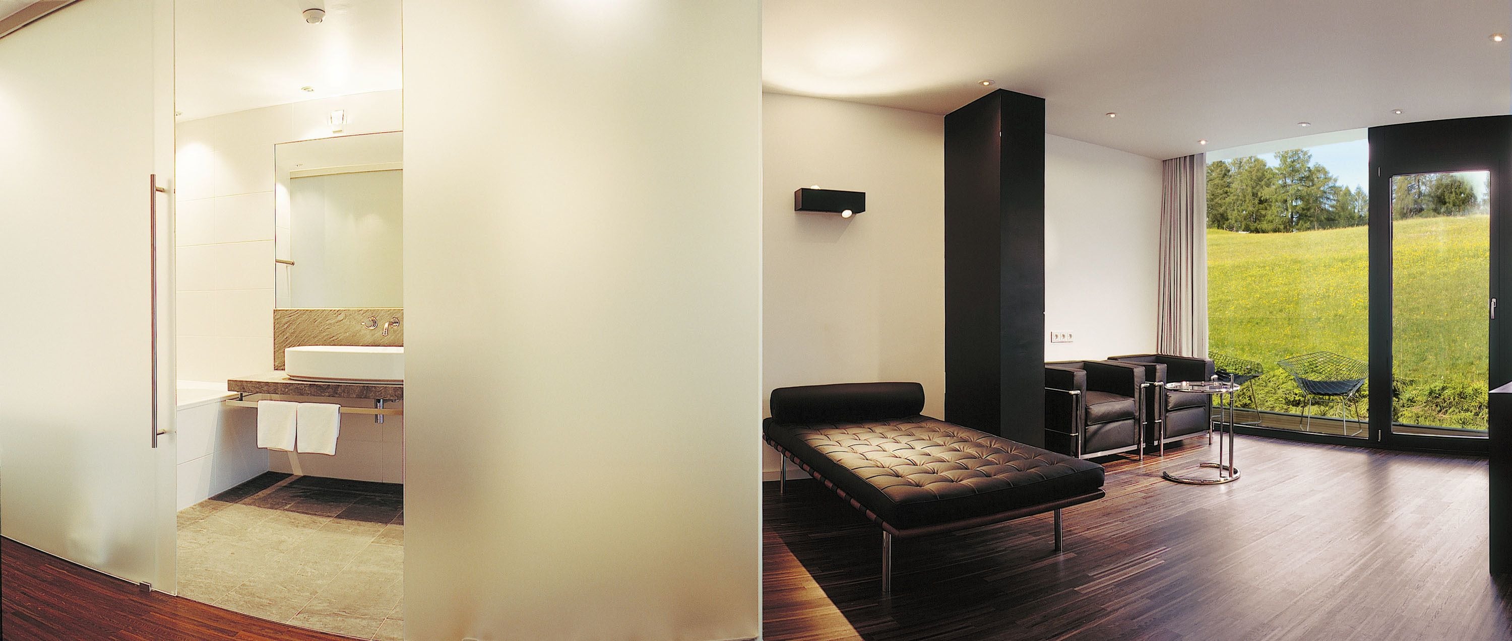 A modern bathroom with a toilet, sink, and towel rack. The room has a large sliding glass door leading to a balcony with a view of a grassy field.