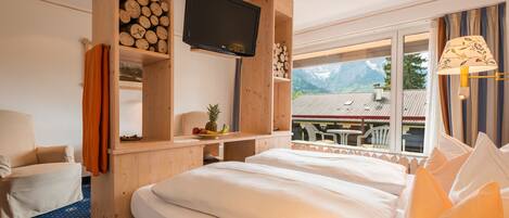 Habitación de hotel con una cama grande, un televisor montado en un estante de madera, una mesita con fruta y vistas a las montañas a través de la ventana.