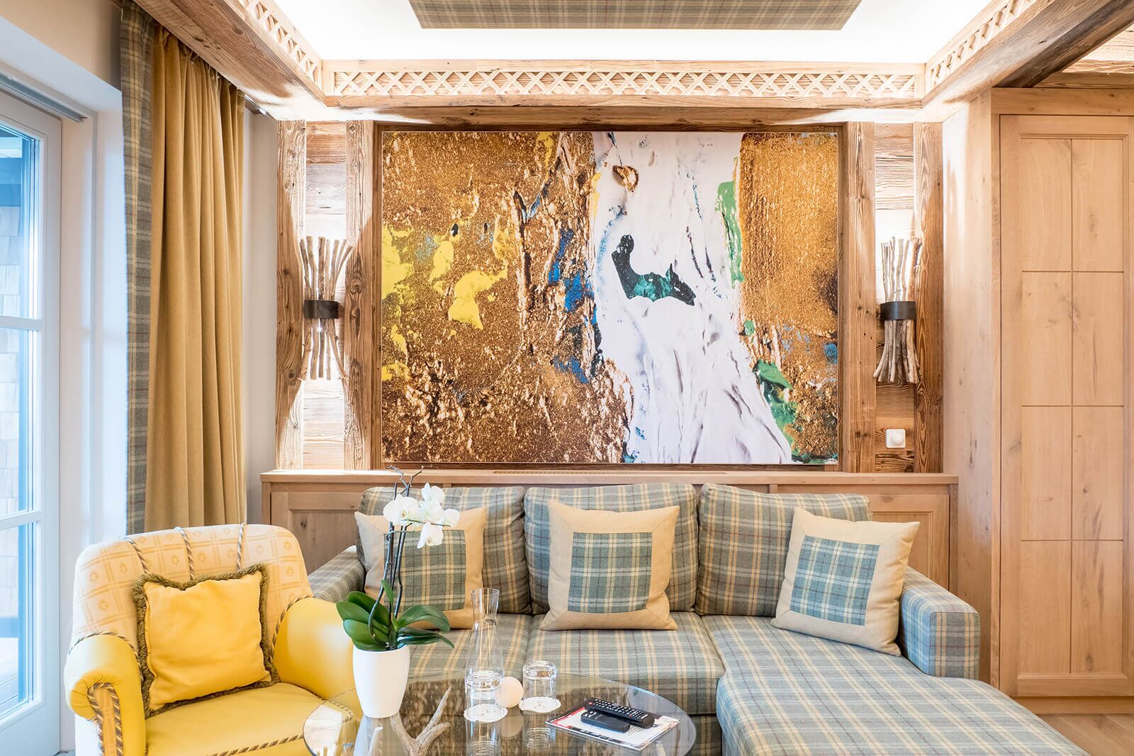 A cozy living room with a large abstract painting, a yellow armchair, a plaid sofa, and a small table with a vase of flowers.
