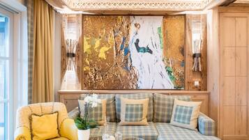 A cozy living room with a large abstract painting, a yellow armchair, a plaid sofa, and a small table with a vase of flowers.