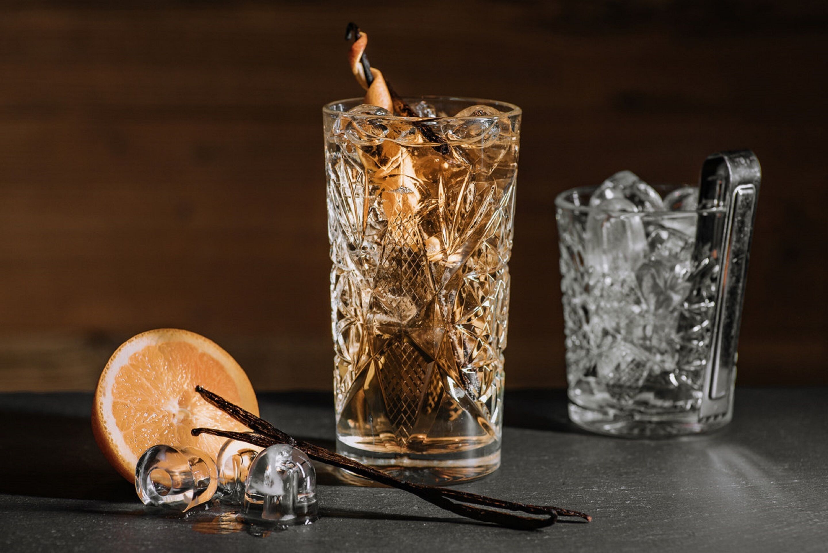 A tall, textured glass with ice cubes, a slice of orange, and a vanilla pod.