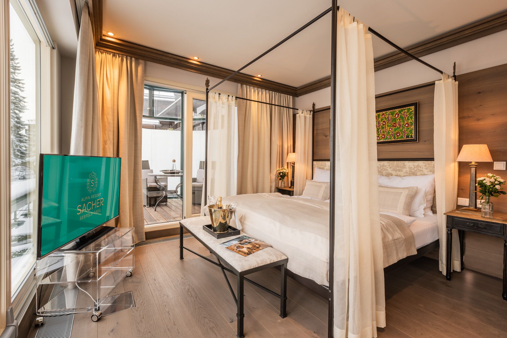 A hotel room with a four-poster bed, a television, a bedside table with a magazine, a nightstand with a lamp, and a view of the outdoors.