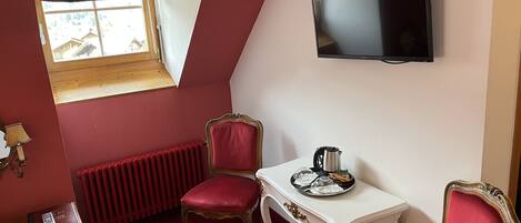 A room with a red carpet, a white table with a kettle, two red chairs, a wooden dresser, a radiator, and a TV on the wall.