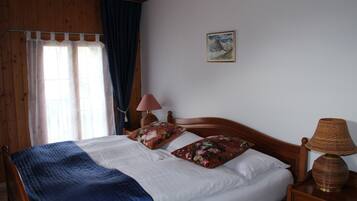 A bedroom with a wooden bed, white bedding, a blue blanket, a wooden nightstand with a lamp, and a painting on the wall.