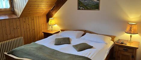 A cozy bedroom with a wooden bed, two bedside tables with lamps, a radiator, and a mountain painting on the wall.