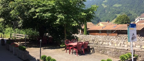 An outdoor seating area with stone walls and red chairs, surrounded by greenery and a mountain backdrop.
