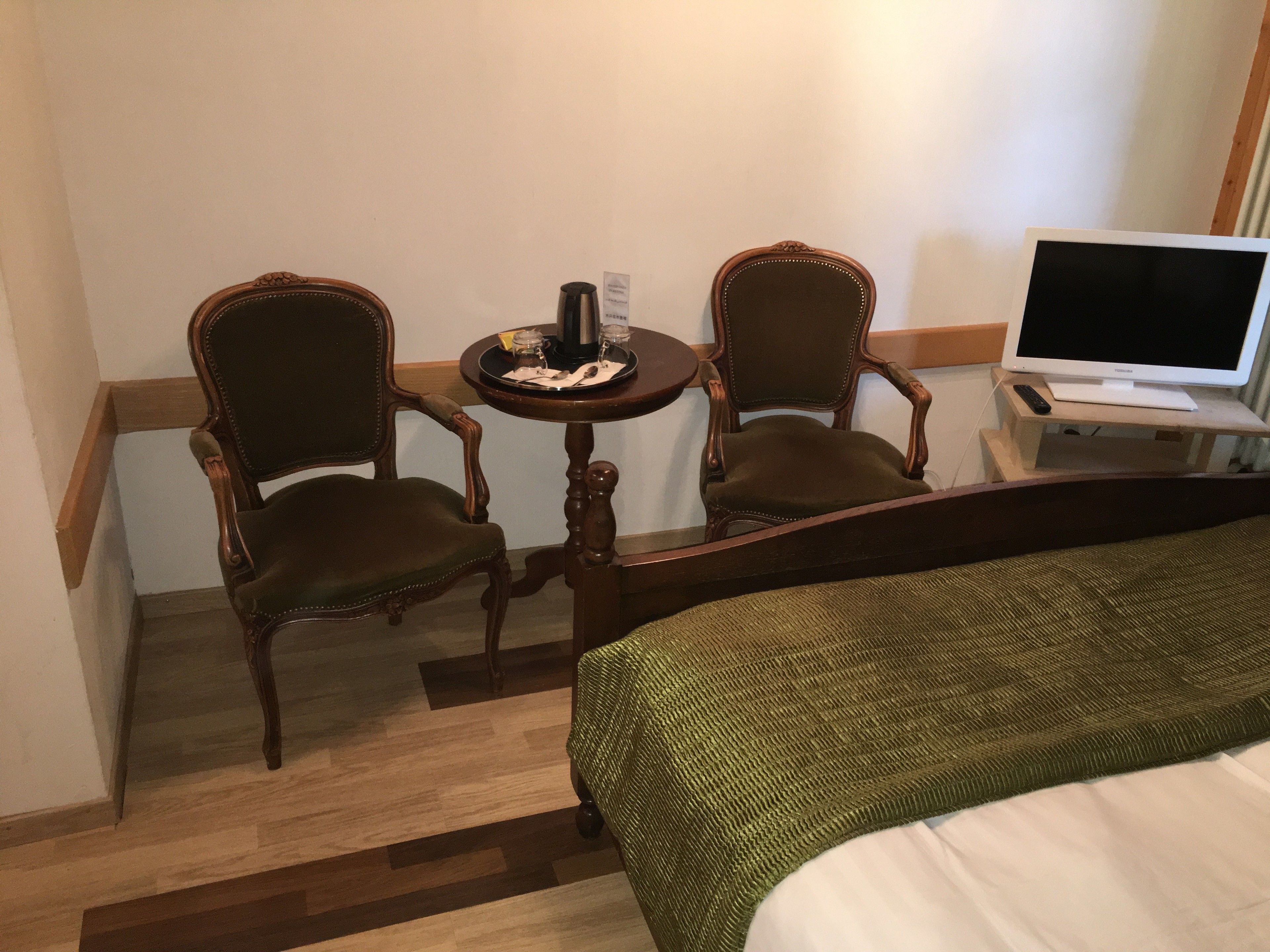 Habitación de hotel con cama, dos sillas de madera, una mesita redonda con bandeja y televisor sobre un soporte.