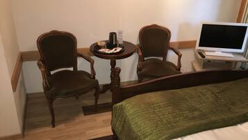 Habitación de hotel con cama, dos sillas de madera, una mesita redonda con bandeja y televisor sobre un soporte.