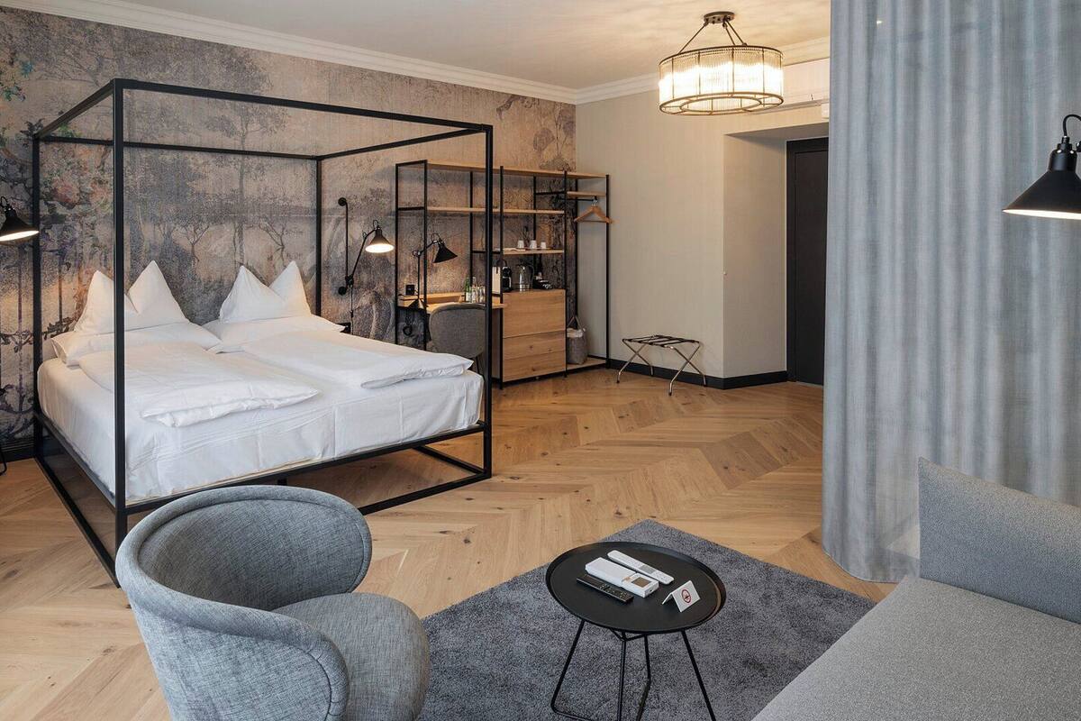 Une chambre à coucher moderne avec un lit à baldaquin, un fauteuil gris, une petite table ronde et un meuble avec miroir.