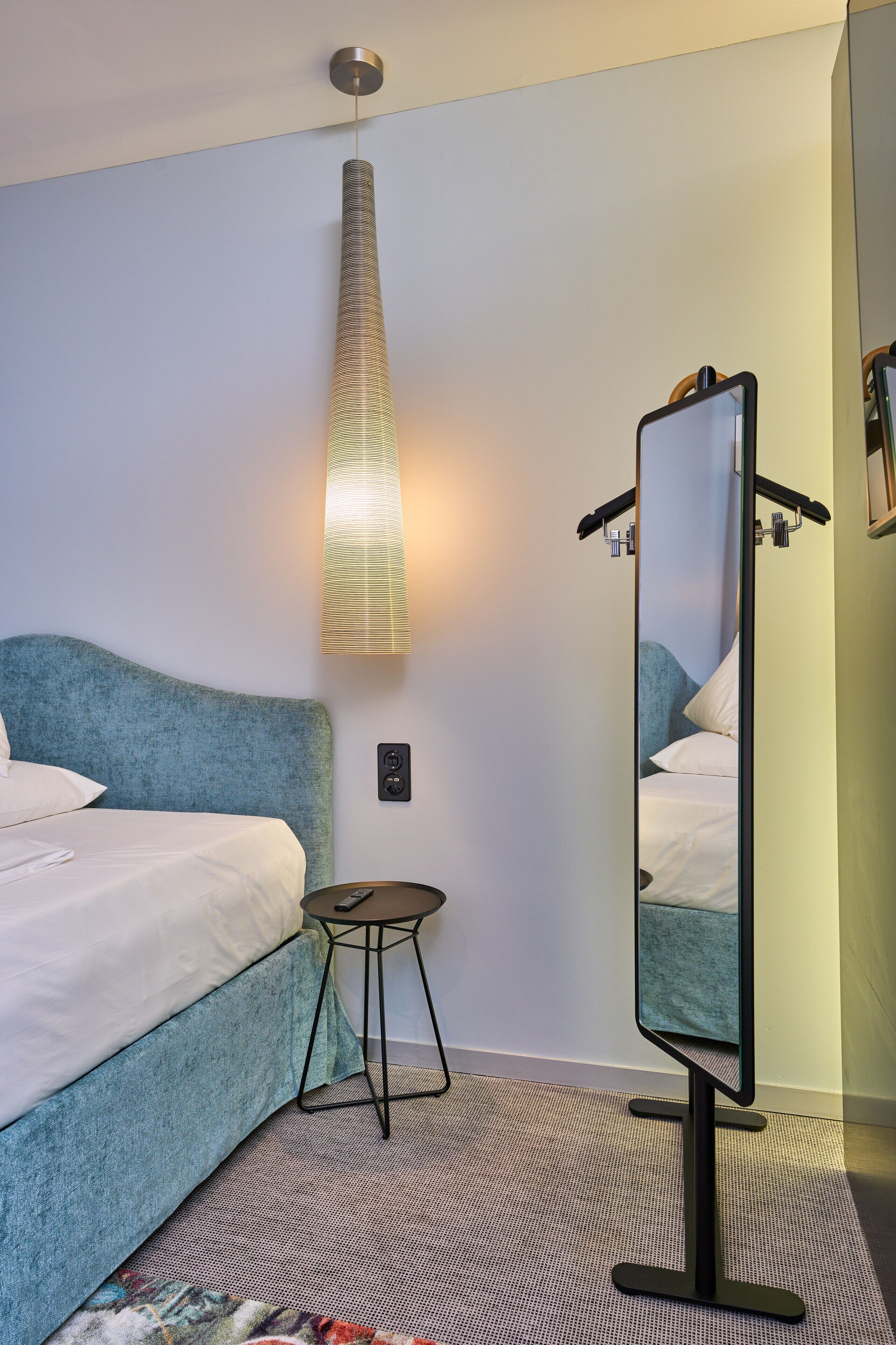 A hotel room with a bed, a bedside table, a mirror, and a wall-mounted light fixture.