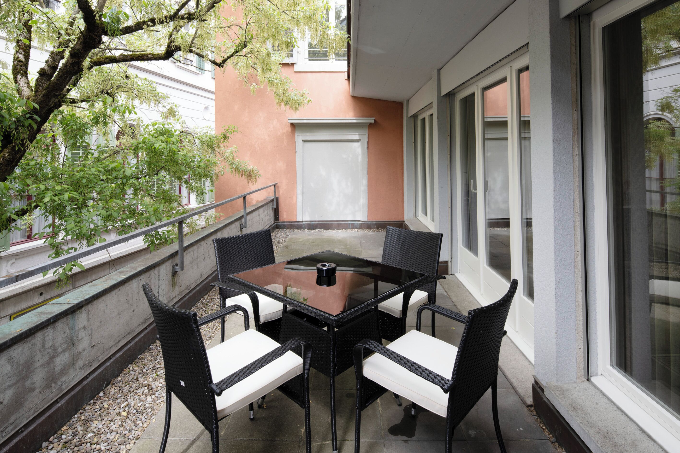 Ein Balkon mit einem Glastisch und vier Stühlen, eine weiße Tür und ein Gebäude mit Fenstern im Hintergrund.