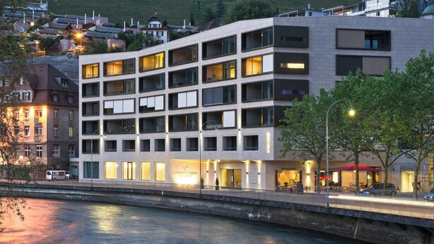 A multi-story hotel with a modern design, located near a river with a bridge.