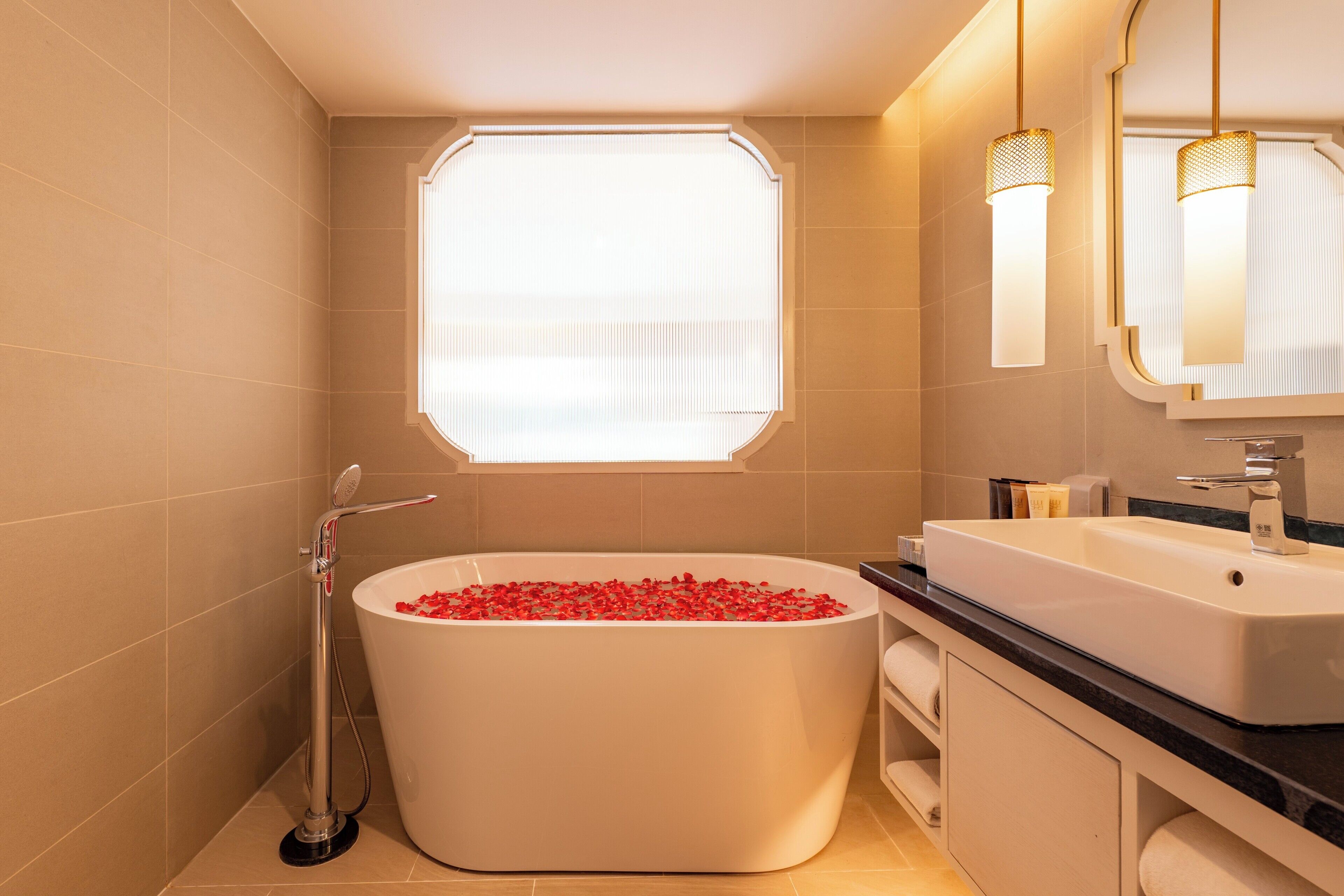 A modern bathroom with a freestanding bathtub filled with red bath beads, a large window, and a vanity with a mirror.