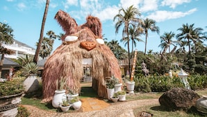 A large thatched structure resembling a creature's face with a pathway leading to it, surrounded by potted plants and palm trees.