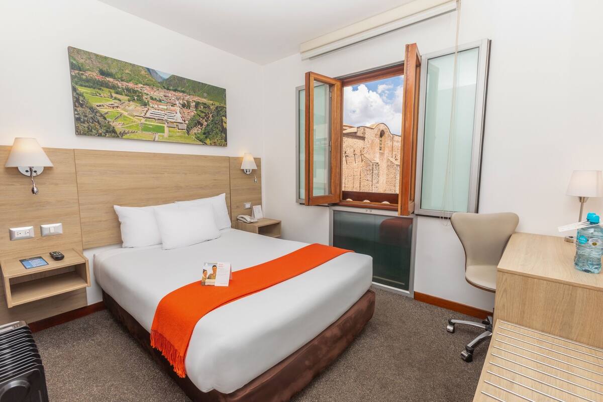 Habitación de hotel con una cama grande, un escritorio con silla, una mesita de noche y una ventana con vista a un edificio.
