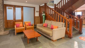 Un vestíbulo con una escalera de madera, un sofá beige con cojines naranjas, una mesa de centro de madera y un extintor.
