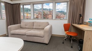 Habitación de hotel con sofá, sillón, escritorio y vistas a un paisaje urbano a través de amplias ventanas.