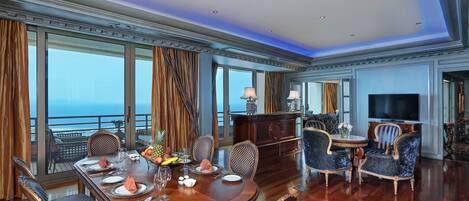 Un comedor lujoso con una gran mesa de madera dispuesta para seis comensales, rodeada de sillas ornamentadas. Se ve un televisor y una mesita con un jarrón de flores al fondo.
