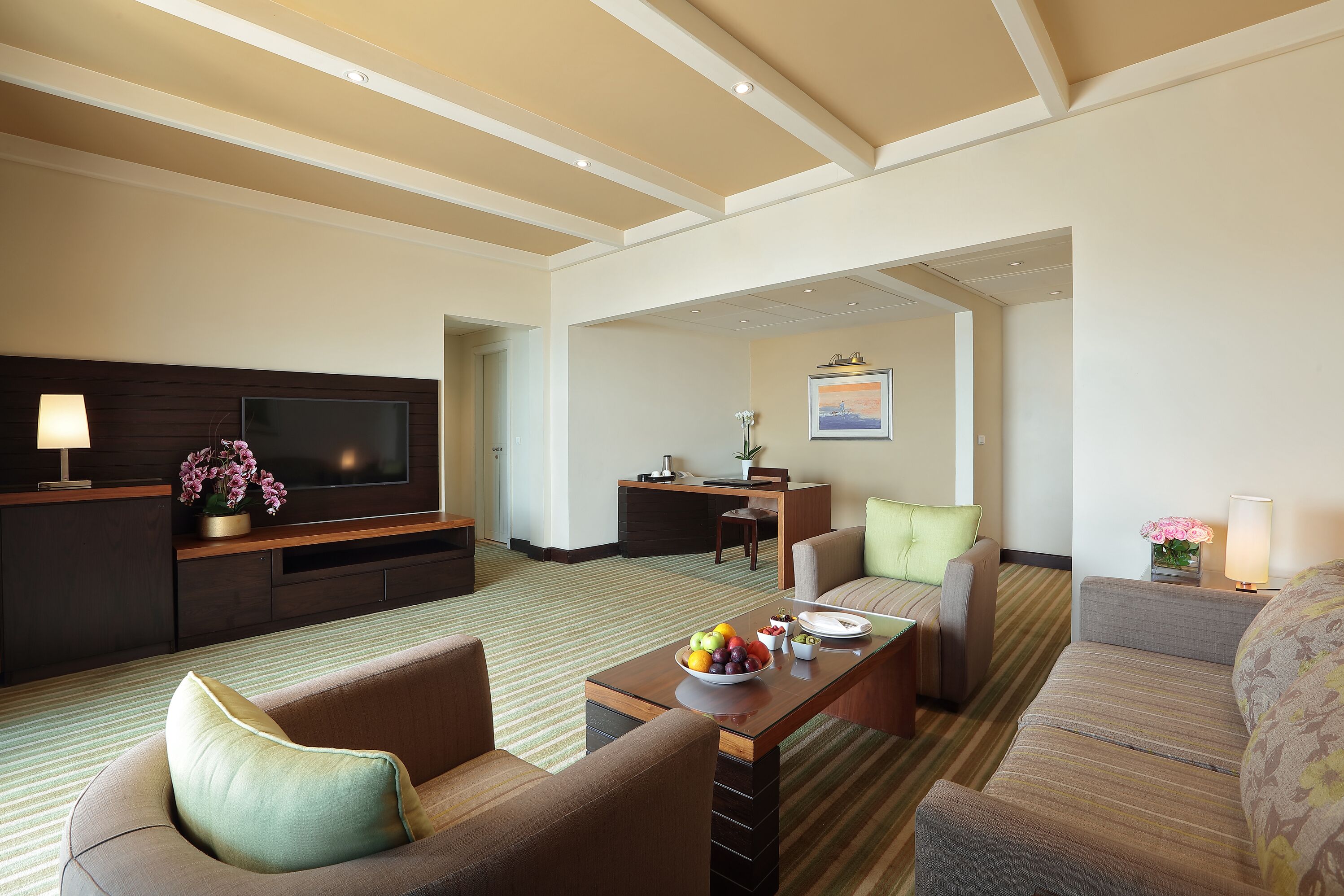 A hotel room with a flat-screen TV, a dining table with fruit, a sofa, and a desk with a lamp.