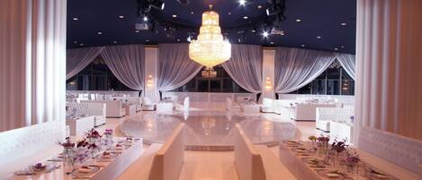 A spacious, elegantly decorated banquet hall with a central chandelier, white drapery, and plush seating arrangements.