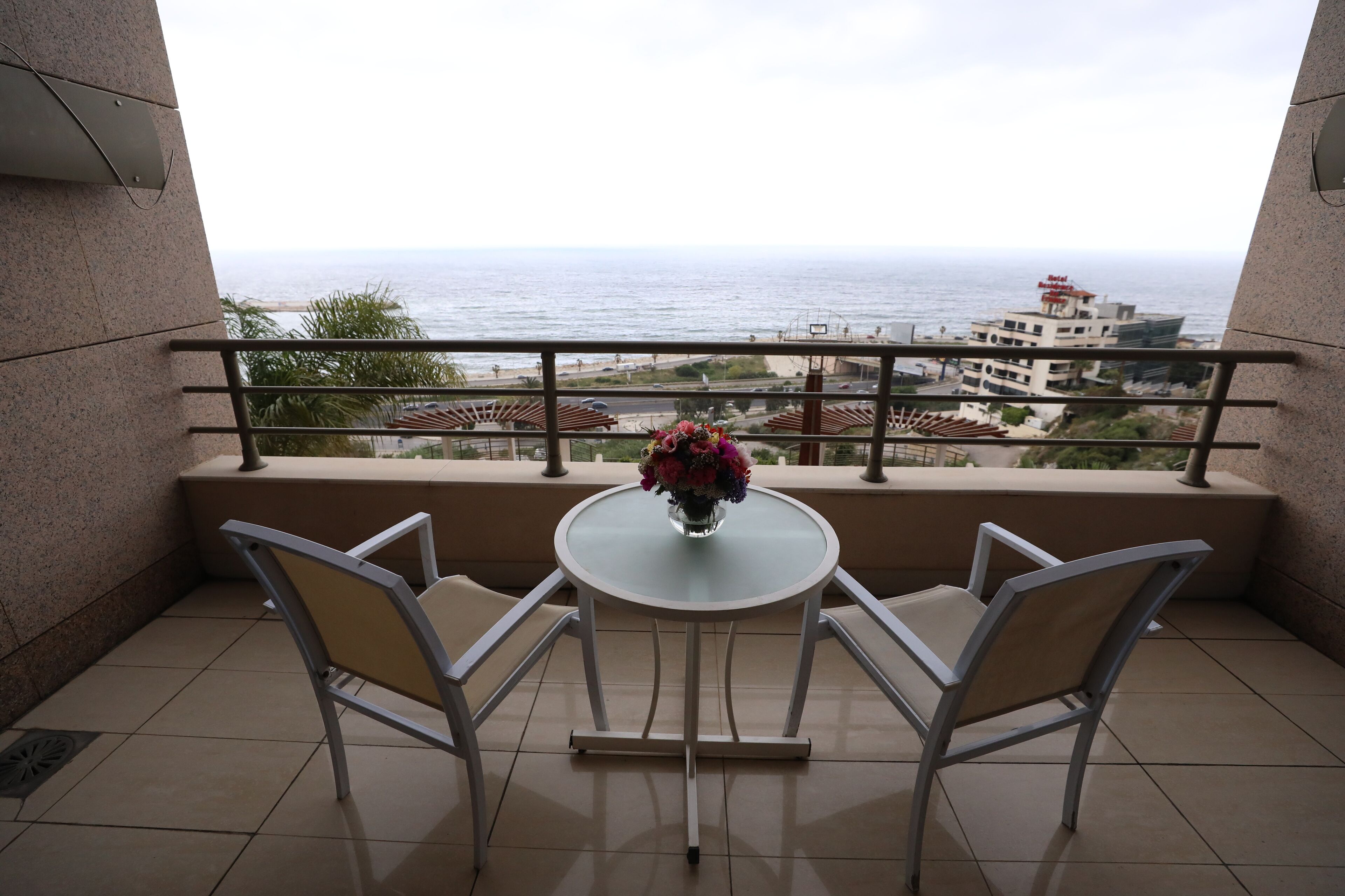 A balcony with a table and chairs, a vase with flowers, and a view of the sea and buildings.